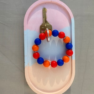 May include: A key ring with a colorful beaded bracelet in blue, orange, and red. The key ring is hanging on a key hook in a pink and blue concrete dish.