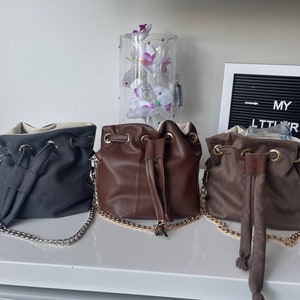 Könnte beinhalten: Drei Kordel-Eimer-Taschen mit Kettenriemen. Die Taschen sind grau, braun und beige. Die Taschen sind auf einer weißen Oberfläche aufgereiht.