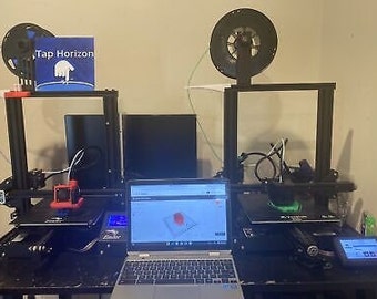 May include: Two black 3D printers with a laptop computer in the middle. The printers have spools of filament and are set up on a black surface. The laptop screen shows a 3D model of a design.