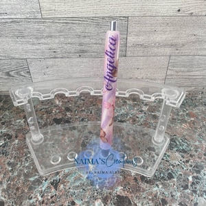 May include: A clear acrylic pen holder with a pen. The pen is light purple with a floral design and the name "Angelica" in purple script. The pen holder has a curved design and the text "NAIMA'S Creations" is visible.