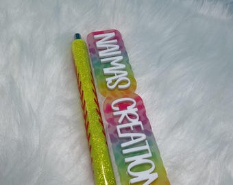 May include: A yellow glitter pen with red stitching details resembling a softball, paired with a rectangular resin tag featuring a multicolored leopard print pattern and the white text NAIMAS CREATIONS displayed in a vertical orientation.