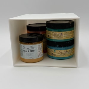 May include: A white box contains several jars of chalk paint. One jar is labeled "Annie Sloan CHALK PAINT" in Arles color. Two jars of "Dixie Belle" chalk mineral paint are stacked, labeled "Mermaid Tail" and "Pure Ocean".