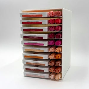 May include: A white, multi-tiered organizer filled with colorful ink pads and markers. The labels read: Calypso Coral, Poppy Parade, Real Red, Cherry Cobbler, Magenta Madness, Pale Papaya, Pumpkin Pie, Cinnamon Cider, and Cajun Craze.
