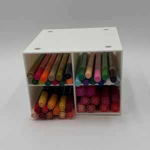 May include: A white, four-compartment organizer filled with various colored acrylic paint pens. The pens are arranged by color, including shades of pink, orange, green, and brown. The organizer is made of a solid white material.