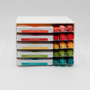 May include: A white plastic storage organizer with a rainbow of colored markers. The labels on the organizer read "Calypso Coral", "Poppy Parade", "So Saffron", "Granny Apple Green", and "Bermuda Bay".