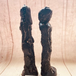 Può includere: Due candele nere, alte e sottili, con una superficie ruvida e testurizzata. Le candele sono in piedi su una superficie di legno chiaro. Il testo "SHUN DESIGNS" è visibile su un'etichetta bianca al centro inferiore dell'immagine.
