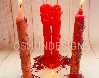 Puede incluir: Tres velas rojas, dos altas y una pequeña, están dispuestas sobre una superficie de madera. Las velas altas están decoradas con hierbas y la vela pequeña está en un recipiente de vidrio. Pétalos de rosa roja están esparcidos alrededor de las velas.