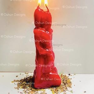 Puede incluir: Una vela roja con dos llamas brillantes. La vela tiene una superficie texturizada y se encuentra sobre un lecho de hierbas y hojas secas. La imagen incluye el texto "Oshun Designs" y "Oshundesigns.org".