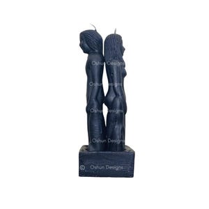 Peut inclure: Une bougie noire représentant deux figures dos à dos. Les figures sont sculptées avec des détails et se tiennent sur une base rectangulaire. La base porte l'inscription "Oshun Designs".