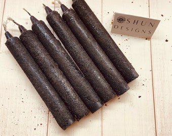 May include: Seven black taper candles with a textured, rough surface. The candles are arranged in a fan shape on a light wood surface. The text "SHUN DESIGNS" is visible on a small card in the upper right corner of the image.