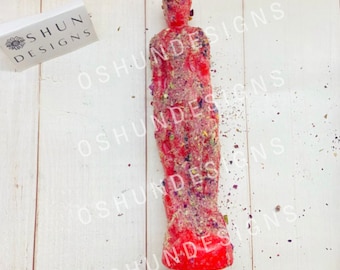 May include: A red figure candle, adorned with dried herbs and flowers, stands upright. The candle is in the shape of a human figure. A white box with the text "SHUN DESIGNS" is in the background.
