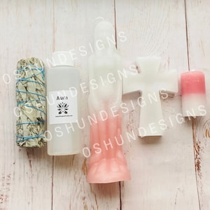 May include: A set of four items for spiritual cleansing and energy work. The set includes a white sage smudge stick, a clear bottle of aura cleansing spray, a white cross candle, and a pink and white pillar candle. The aura cleansing spray bottle has a label that reads "Aura".