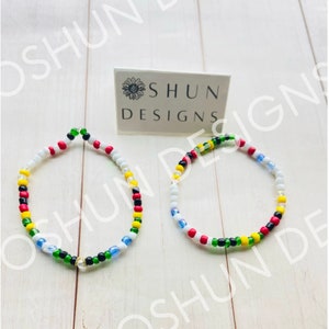 May include: Two colourful beaded bracelets with a white, red, yellow, green and black colour scheme. The bracelets are on a white wooden surface with a business card that says "Shun Designs".