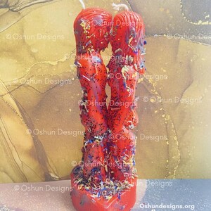 May include: A red candle in the shape of two figures embracing, decorated with glitter and dried flowers. The candle sits on a heart-shaped base. The text "Oshun Designs" is visible on the image.
