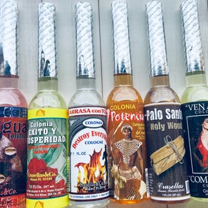 Puede incluir: Seis botellas de vidrio de colonia con etiquetas coloridas y tapas plateadas. Las etiquetas presentan imágenes y texto en español. Las botellas están dispuestas en una fila sobre una superficie de madera blanca. Las colonias son: Eggua, Colonia Exito y Prosperidad, Colonia Destroy Every Cologne, Colonia Potencia, Palo Santo Holy Wood y Ven a Mi Colonia.