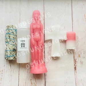 May include: An assortment of spiritual items on a light wood surface. Includes a pink goddess candle, a white angel mold, a bundle of sage, a bottle labeled "Aura", and a two-tone pink and white candle.
