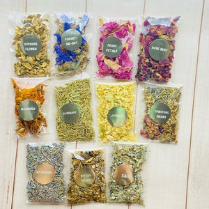 Puede incluir: Una colección de diez pequeñas bolsas de plástico transparente llenas de hierbas y flores secas. Cada bolsa tiene una etiqueta dorada con texto, incluyendo "Sophore Flower", "Forget-Me-Not", "Peony Petals" y "Rose Buds". Otras etiquetas dicen "Marigold", "Rosemary", "Jasmine", "Spiritual Herbs", "Lavender", "Witchy Herbs" y "Sage".