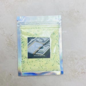 May include: A clear, holographic zip-lock bag filled with a pale green substance and small green particles. A rectangular label shows the words "Money/Dinero" above a money graphic. The website Oshundesigns.org is also visible.