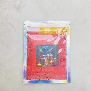 May include: A small, clear, resealable pouch filled with red granules. A label on the pouch reads "Love Spell / Hechizo De Amor" with a design of candles and bottles. The pouch has a holographic sheen.