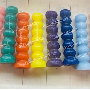 May include: A collection of colorful, pillar-style candles in various shades, including white, pink, teal, orange, yellow, purple, blue, red, and black. Each candle has a unique, stacked design and a single wick. The brand name "OSHUNDESIGNS" is visible.