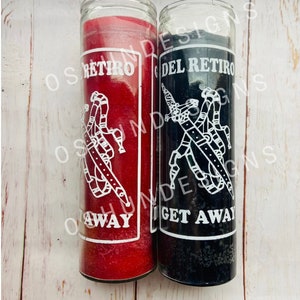 May include: Two glass candle jars with black and red wax. The jars have white labels with the text "Del Retiro" and "Get Away" on the black jar and "Retiro" and "Away" on the red jar. The labels also feature a graphic of a hand holding a sword.