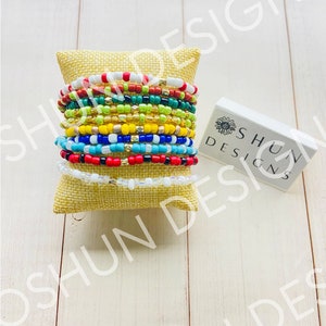 May include: A collection of beaded bracelets in various colors, including red, white, blue, green, yellow, and gold. The bracelets are stacked on a small, square burlap pillow. A white box with the text "SHUN DESIGNS" is to the right.