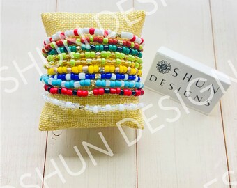May include: A collection of beaded bracelets in various colors, including red, white, blue, green, yellow, and gold. The bracelets are stacked on a small, square burlap pillow. A white box with the text "SHUN DESIGNS" is to the right.