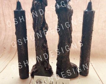 May include: Three black candles, two are tall and twisted, one is shorter and straight. The candles are on a light wood surface. The text "OSHUN DESIGNS" is on a white label.
