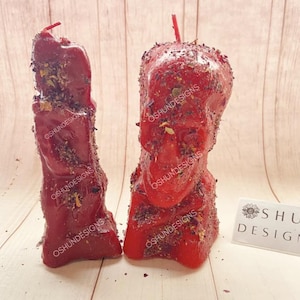 May include: Two red skull-shaped candles with dried herbs and flowers embedded in the wax. The candles are on a wooden surface. The text "OSHUNDESIGNS" is visible on the candles.