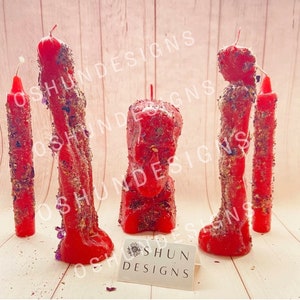 May include: Three red candles with herbs and crystals embedded in them. The candles are in the shape of a person, a head, and a long thin candle. The candles are on a light wood surface. The text "Shun Designs" is on a white card in the bottom right corner of the image.