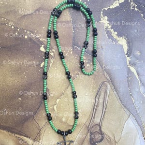 May include: A necklace made of green and black beads, with a silver-coloured pendant depicting a bow and arrow. The beads are strung on a cord. The necklace is shown against a textured background.