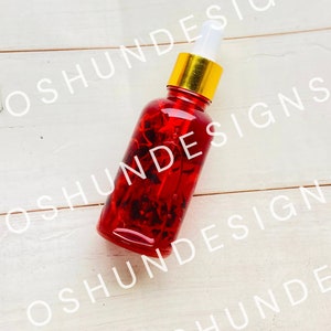 Puede incluir: Una botella de vidrio roja con tapa dorada y gotero. La botella está llena de un líquido rojo y tiene pequeñas flores rojas en su interior.