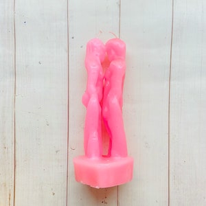 May include: Pink candle shaped like two figures embracing. The candle is on a white wooden surface.