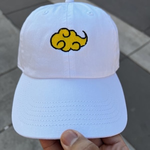 May include: White baseball cap with a yellow cloud design outlined in black. The cap has a curved brim and a fabric strap closure. The cloud design is embroidered on the front of the cap.