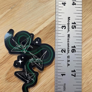 May include: A sticker featuring a green snake with origami paper cranes in a black and white design. The snake is curled around the cranes, and there are small white stars in the background.