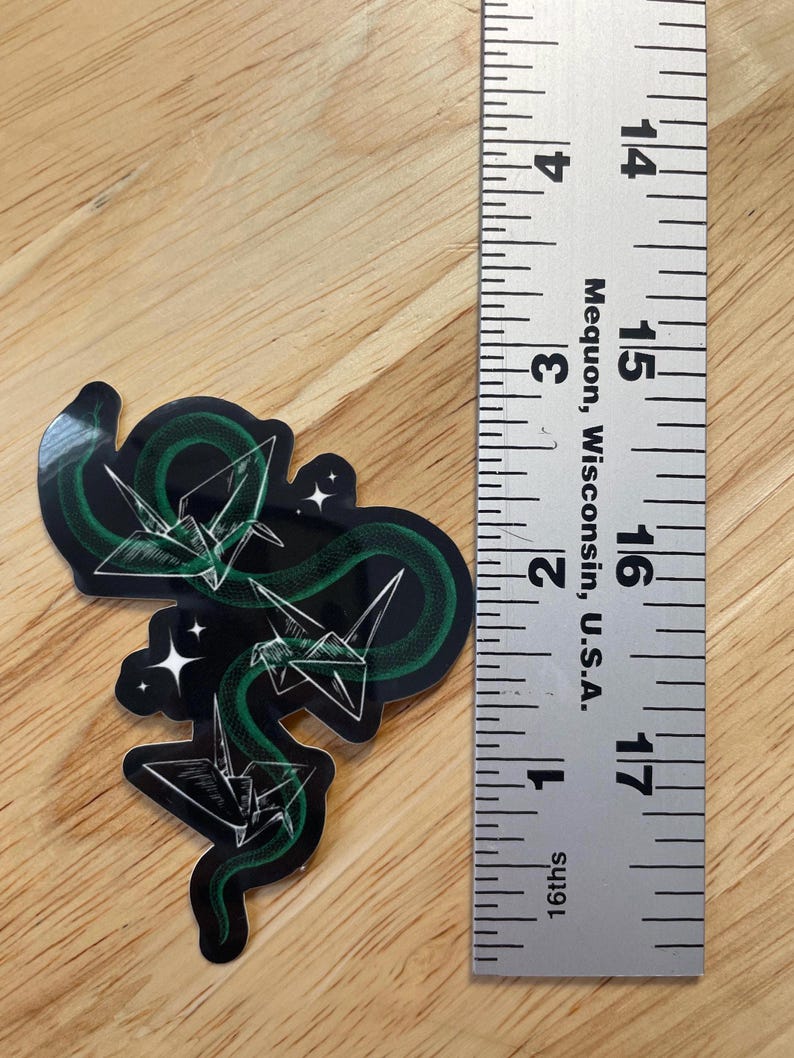 May include: A sticker featuring a green snake with origami paper cranes in a black and white design. The snake is curled around the cranes, and there are small white stars in the background.