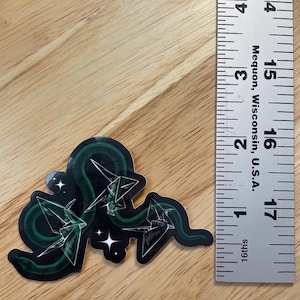 May include: A green snake with a black background and white origami cranes. The snake is curled around the cranes and there are white stars in the background.