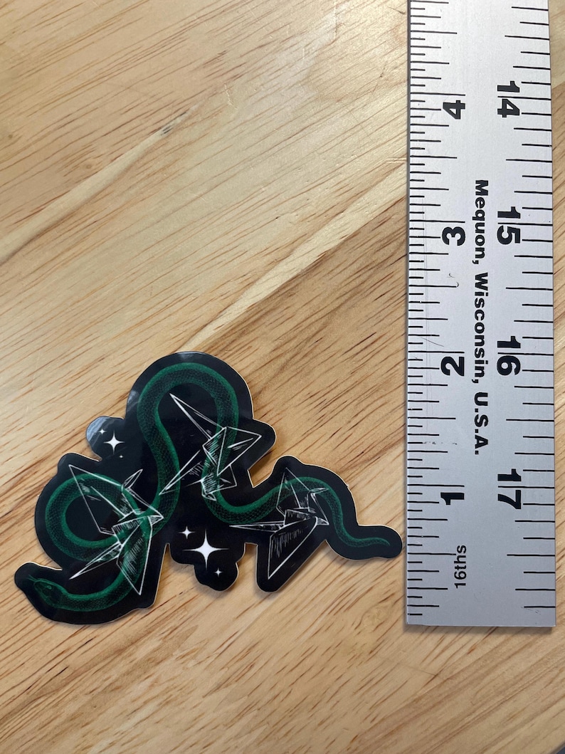 May include: A green snake with a black background and white origami cranes. The snake is curled around the cranes and there are white stars in the background.