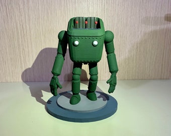 Puede incluir: Figura de robot verde con cabeza en forma de caja y extremidades articuladas. El robot tiene dos ojos circulares blancos y varillas rojas en la parte superior. Se apoya sobre una base circular gris y azul, con un diseño retro-futurista.