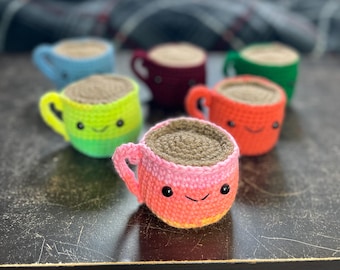 May include: A collection of six crocheted coffee mugs with smiling faces. The mugs are in various colors, including green, red, pink, blue, and brown.