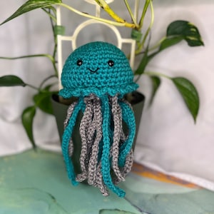 May include: A teal crocheted jellyfish with a smiling face and gray tentacles. The jellyfish is sitting on a pot with a green plant.
