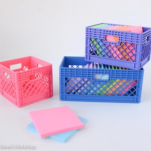 May include: Three plastic storage crates in pink, blue, and purple, featuring a lattice design and integrated handles. The blue and purple crates are stacked and filled with colorful square sticky notes, while a pink and blue sticky note lie flat.