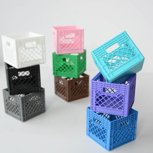 May include: A collection of small, colorful plastic crates in various colors, including white, black, gray, pink, green, brown, teal, purple, and blue. The crates are stacked in small towers, showcasing their design and color variations.
