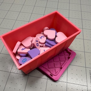 May include: A pink rectangular container filled with heart-shaped and rectangular tokens. The tokens are pink and purple, some with numbers. A purple tray with a geometric pattern is beneath the container.