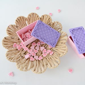 May include: A woven, flower-shaped basket holds pink heart-shaped candies with numbers. Two small pink boxes with purple lids are nearby. Scattered around are more heart candies, all on a white surface.
