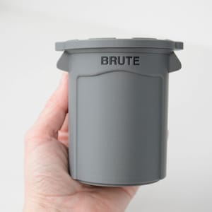 May include: A gray, cylindrical container with the word "BRUTE" printed on the lid. The container has a handle-like design on the sides. The object is held in a hand against a white background.
