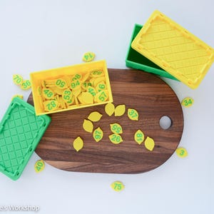 May include: A collection of small yellow lemon-shaped tokens with green printed numbers, contained within a yellow rectangular plastic bin. Additional green and yellow plastic bins with lids are present, alongside loose tokens scattered on a dark wood board.