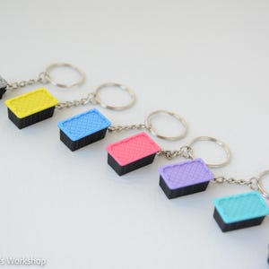 May include: A collection of rectangular keychains, each featuring a black base and a textured lid in various colors including gray, yellow, blue, pink, purple, and light blue, attached to a silver metal split ring by a short chain.