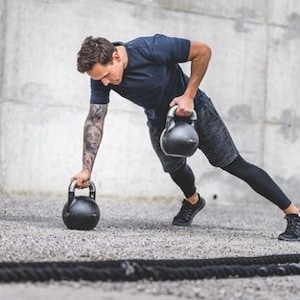 Puede incluir: Un hombre con una camisa y pantalones cortos negros está haciendo un ejercicio de kettlebell. Está sosteniendo una kettlebell en cada mano y está en una posición de estocada baja. Las kettlebells son negras y tienen una superficie texturizada.