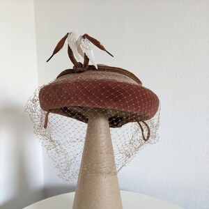 Puede incluir: Un sombrero de pastillero de terciopelo marrón con una decoración de pájaro marrón y blanco y un velo marrón.